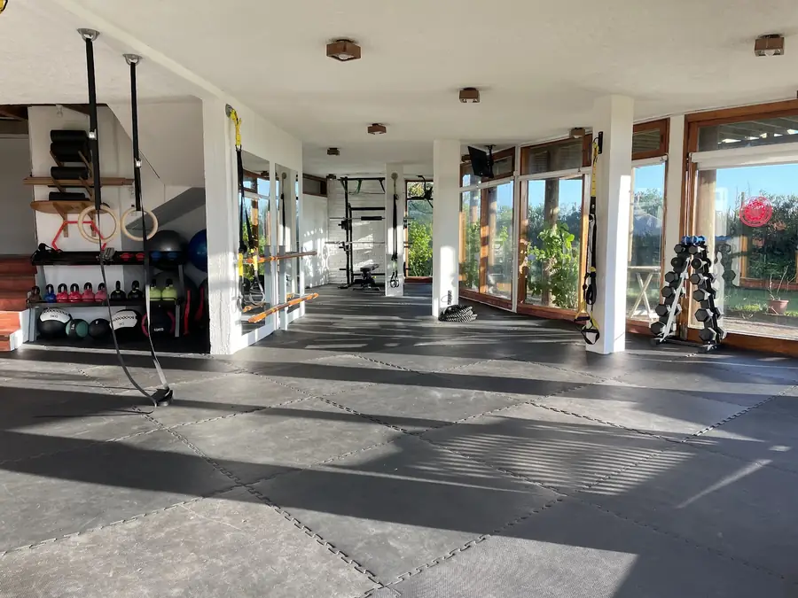 La sala principal de Kai vacía: piso de goma negro, columnas blancas, ventanales con marcos de madera, anillas, pesas rusas y balones